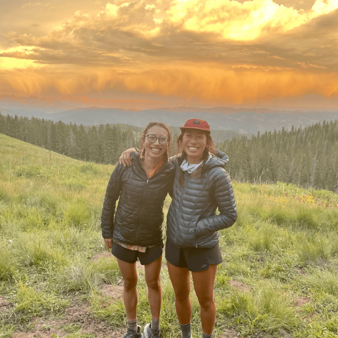 Two women wearing athletic clothing smile in front of a sunset and forest