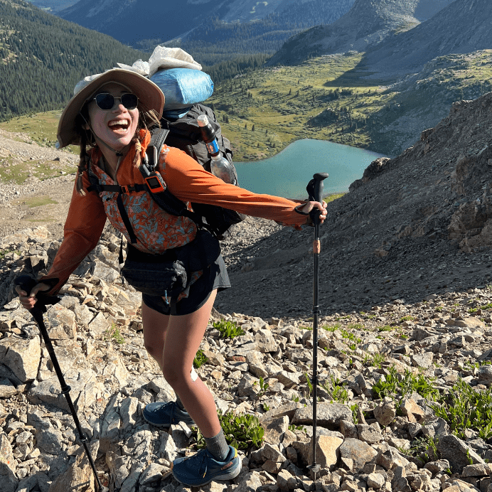 A woman smiles with her mouth open wearing a backpack and hat in front of a lake